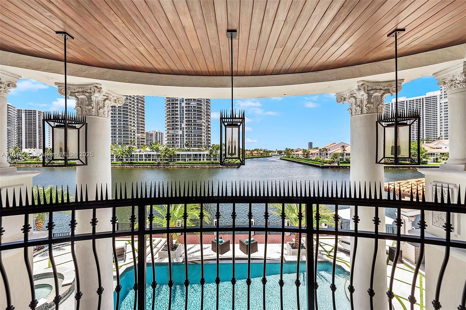 This elevated balcony offers a sophisticated vantage point overlooking a private swimming pool and a tranquil canal. The space is defined by classical columns with ornate capitals, a warm wood-paneled ceiling, and elegant hanging lanterns, creating a luxurious outdoor living area. The wrought-iron railing with decorative spikes provides a secure yet open boundary, framing the expansive waterfront view of the surrounding neighborhood.