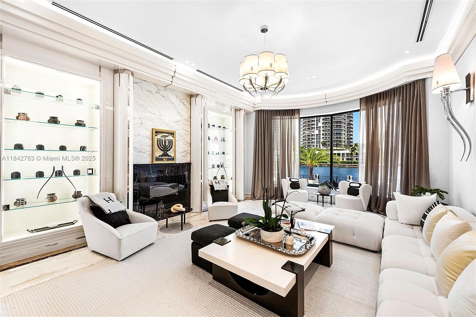 This sophisticated living room features a bright, open-concept layout with a large white sectional sofa and matching armchairs arranged around a central coffee table. A striking stone-clad chimney breast serves as a focal point, flanked by illuminated built-in display shelving that showcases decorative objects. Large windows with elegant drapery frame a scenic waterfront view, while a decorative chandelier adds a touch of luxury to the space.