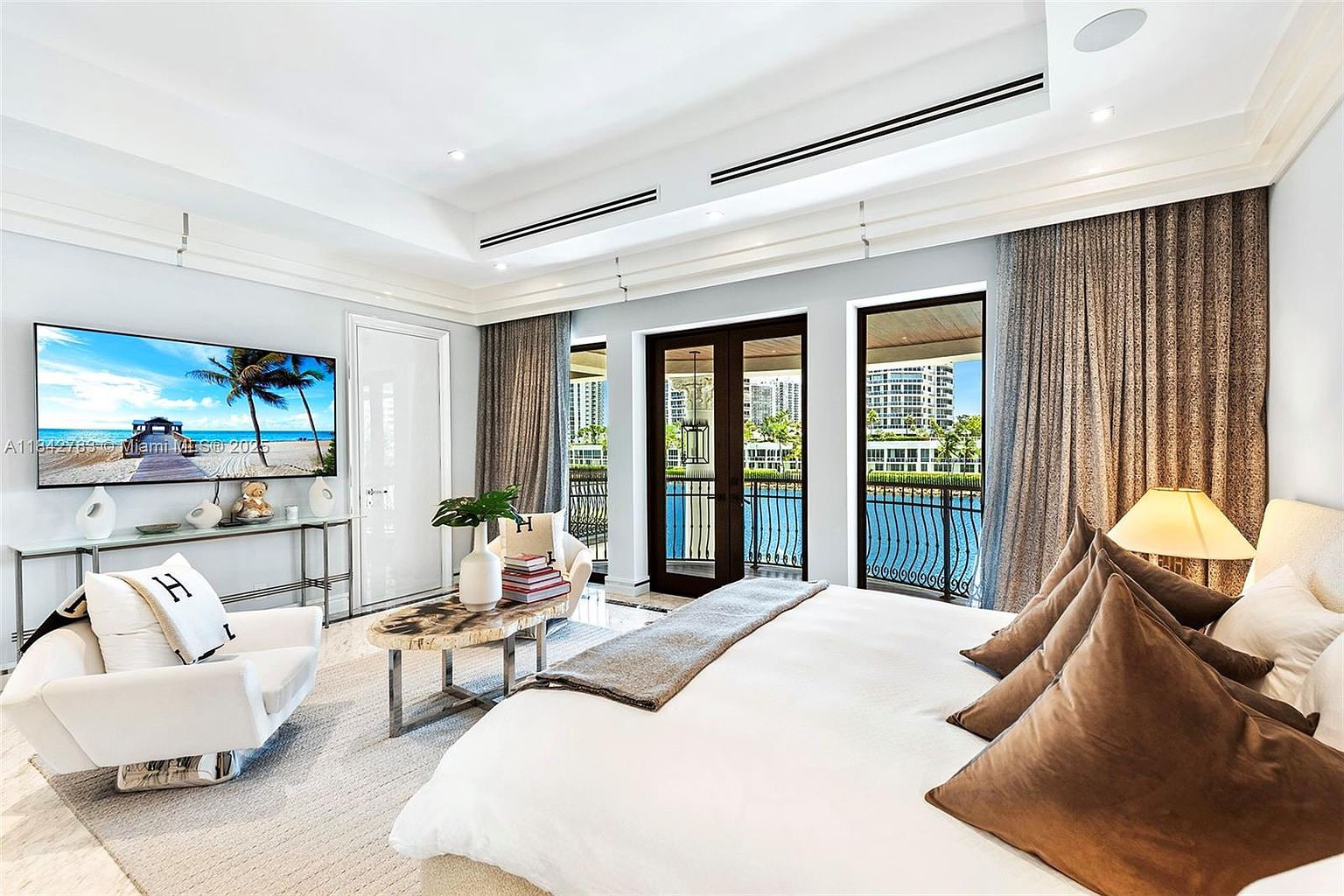 This luxurious primary bedroom features a neutral color palette with plush bedding, brown accent pillows, and a modern white armchair. The room is illuminated by recessed lighting and a bedside lamp, while large glass doors provide access to a balcony with water views. A sleek console table sits beneath a wall-mounted television, creating a sophisticated and serene living space.