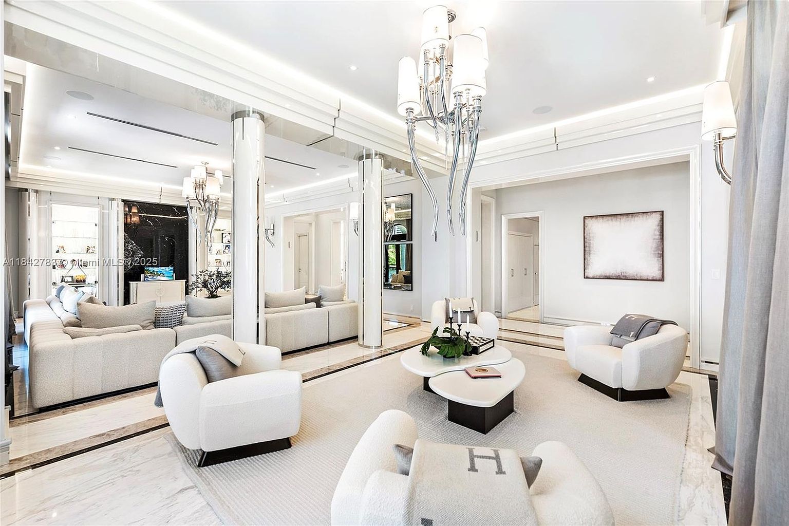 This sophisticated living room features a bright, open-concept design with a neutral color palette and high-end finishes. The space is anchored by a large, plush sectional sofa and several modern armchairs arranged around a unique, multi-tiered coffee table. A striking, sculptural chandelier serves as the focal point, while mirrored columns and reflective surfaces enhance the sense of depth and luxury throughout the room.