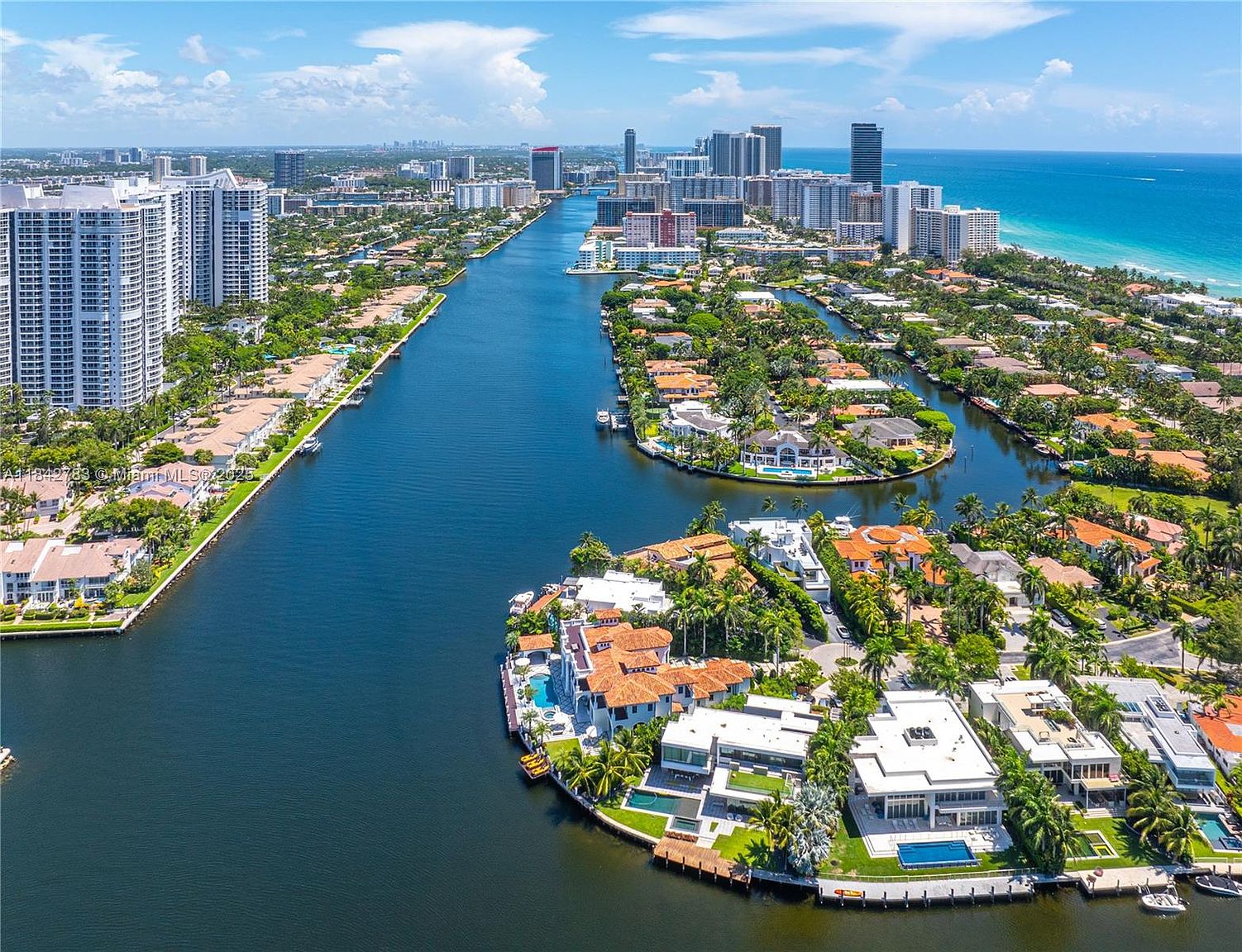 This high-angle aerial view captures a luxurious waterfront neighborhood in Florida, characterized by intricate canal systems and high-end residential estates. The foreground features several modern homes with private pools and docks, while the background showcases a dense skyline of high-rise condominiums meeting the Atlantic Ocean. The perspective emphasizes the prime location, highlighting the seamless integration of coastal living and urban convenience.