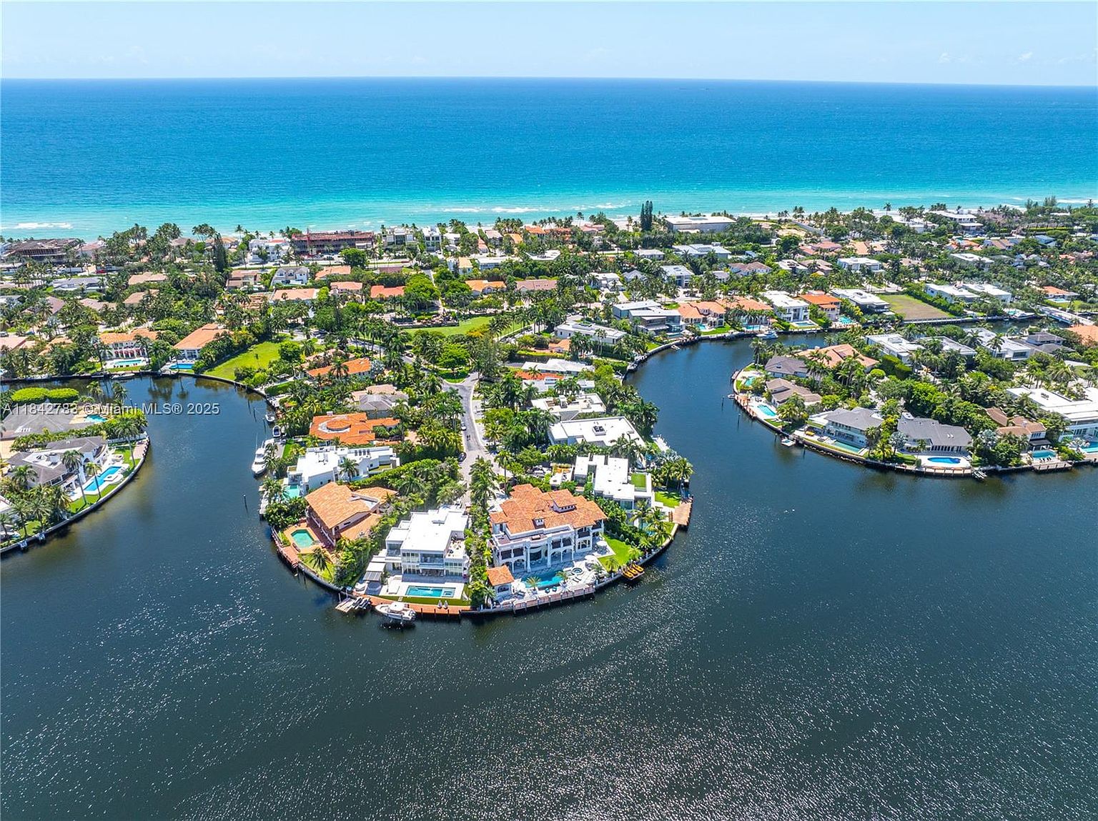 This high-angle aerial view captures a luxurious waterfront residential enclave situated on a peninsula surrounded by calm, deep-blue waterways. The scene features several expansive estates with terracotta-tiled roofs, private swimming pools, and personal boat docks, all set within a lush, tropical landscape. In the background, the turquoise waters of the ocean meet the horizon, emphasizing the prime coastal location of this exclusive neighborhood.