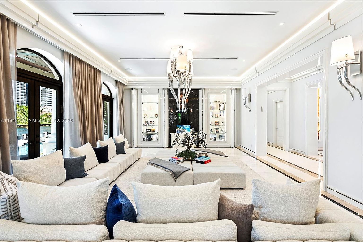 This expansive, high-ceilinged living room features a sophisticated neutral palette with plush, oversized modular seating arranged around a central ottoman. The space is anchored by a striking, multi-tiered crystal chandelier and flanked by symmetrical, illuminated display cabinets that frame a dark, reflective accent wall. Large glass doors with dark frames provide a view of the exterior, while elegant wall sconces and recessed lighting enhance the bright, airy atmosphere.