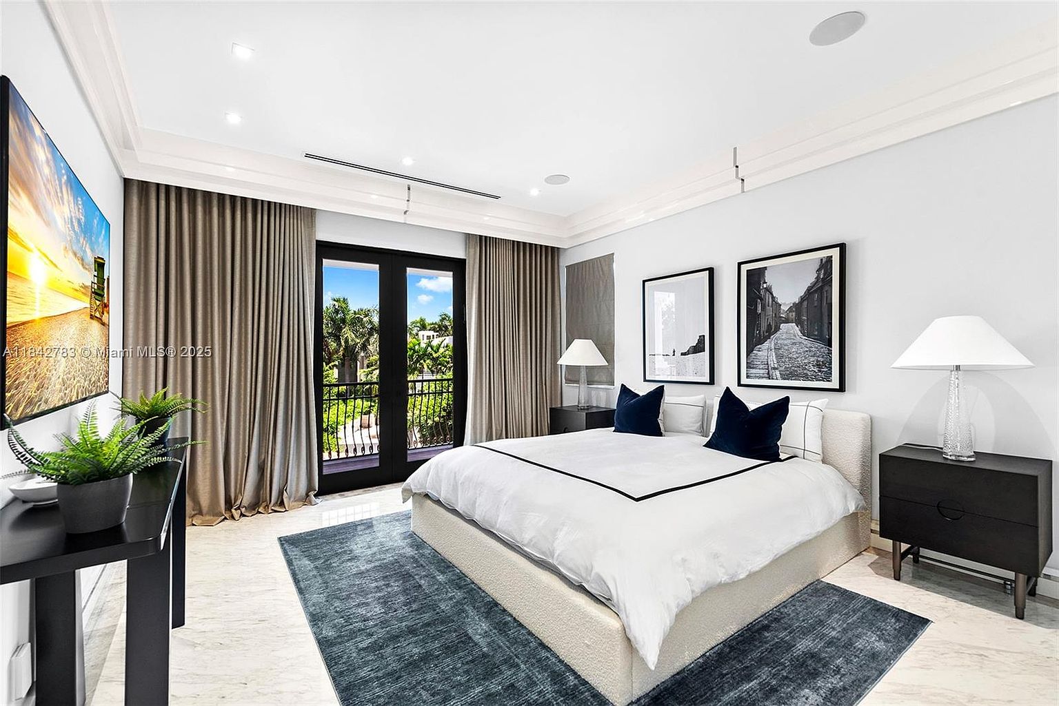 This primary bedroom features a neutral color palette with a large, upholstered bed as the focal point, accented by dark blue throw pillows. The room includes two black nightstands with matching table lamps, framed artwork on the wall, and a large television mounted to the left. A set of double doors with glass panels opens to a balcony, framed by floor-to-ceiling curtains, while a dark area rug adds texture to the light-toned stone flooring.