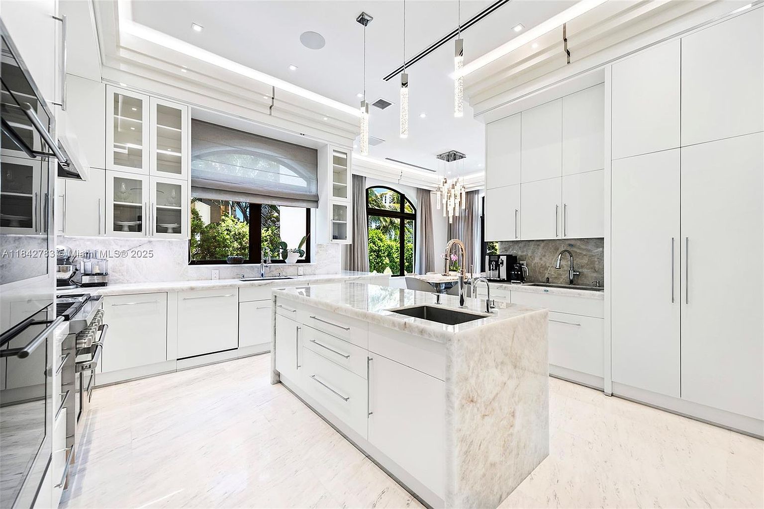 This bright, modern kitchen features crisp white cabinetry, a large central island with a stone countertop, and integrated stainless steel appliances. The space is illuminated by sleek pendant lights and recessed lighting, creating an airy and sophisticated atmosphere. A large window above the sink provides a view of the lush greenery outside, while the neutral color palette and clean lines offer a polished, high-end aesthetic.