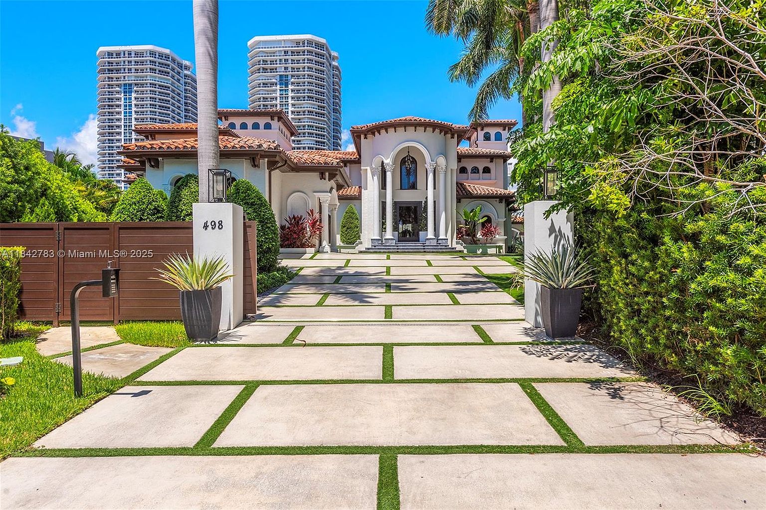 This Mediterranean-style estate features a grand, arched entryway supported by prominent columns and a terracotta-tiled roof. A long, geometric concrete driveway with integrated grass strips leads directly to the front entrance, flanked by two large planters on stone pillars. The property is framed by lush greenery and palm trees, with modern high-rise buildings visible in the background, creating a striking contrast between luxury residential architecture and urban density.