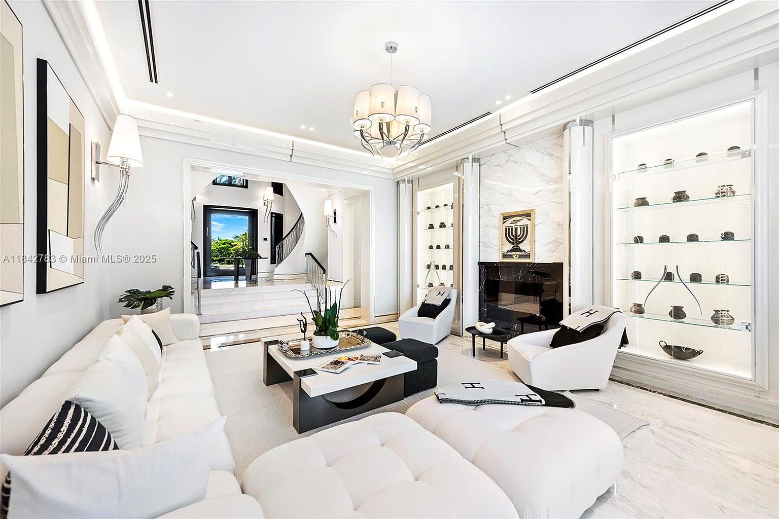 This luxurious living room features a bright, monochromatic palette with plush white seating and a central coffee table. A prominent fireplace with a dark stone surround and a tall marble-clad chimney breast serves as the focal point, flanked by illuminated glass display shelving. The space is accented by a sophisticated chandelier, designer throw blankets, and a clear view through an arched entryway toward a grand staircase.