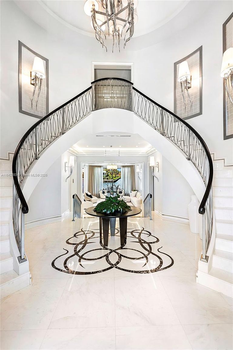This grand foyer features a dramatic, symmetrical curved staircase with intricate metal railings that lead to an upper landing. The space is anchored by a circular center table resting on a decorative, inlaid stone floor pattern, while a large, multi-tiered chandelier hangs from the high ceiling. The bright, white-toned interior is accented by wall sconces and provides a clear, open view through to the living area beyond.