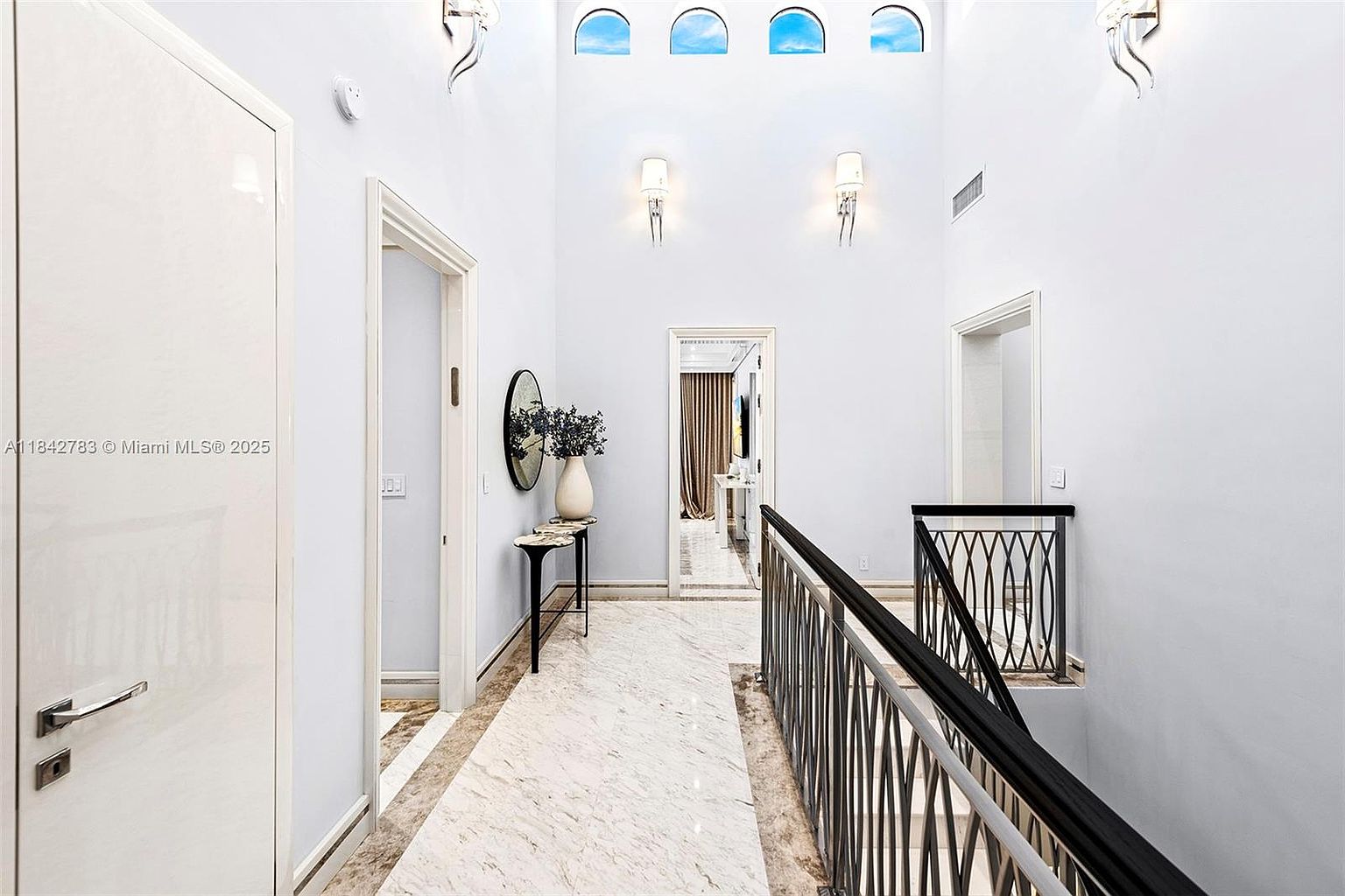 This bright, high-ceilinged hallway features polished light-toned stone flooring and a sleek black metal railing overlooking the staircase. The space is illuminated by wall-mounted sconces and a series of arched windows positioned high on the wall, creating an airy and sophisticated atmosphere. A console table with a decorative mirror and vase adds a touch of elegance to the transition area between rooms.