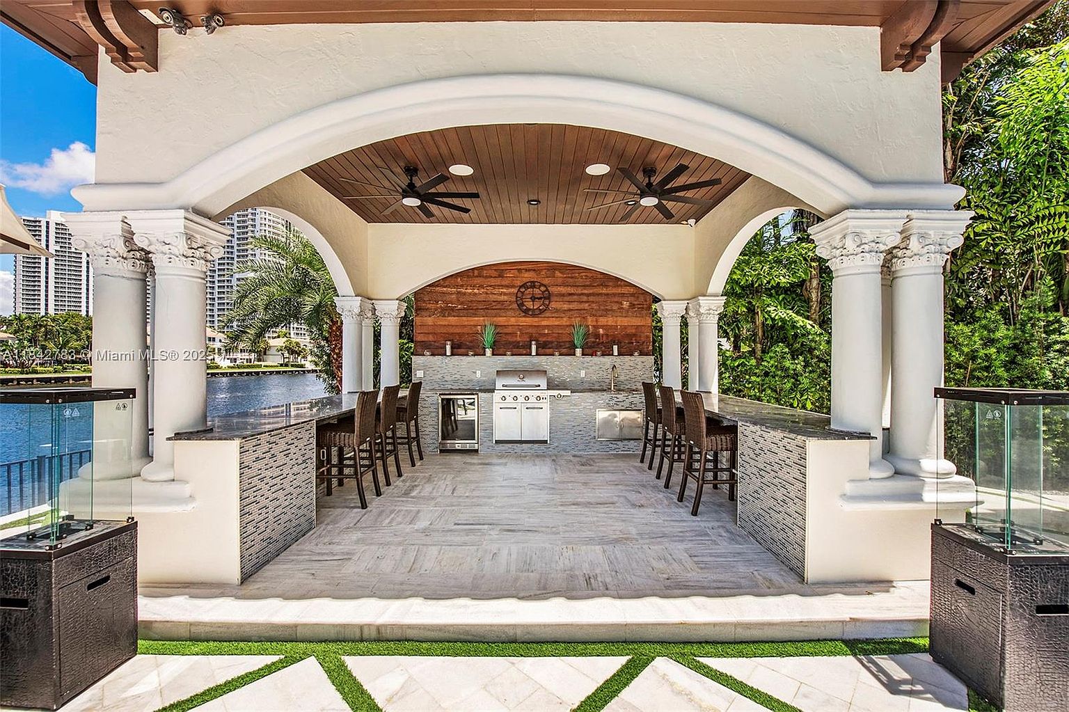 This luxurious outdoor kitchen and lounge area is sheltered by a grand, arched pavilion supported by classical columns. The space features a built-in grill, stainless steel appliances, and a long stone-topped bar with seating, all set against a rustic wood-paneled accent wall. The perspective looks directly through the pavilion toward a waterfront view, creating an elegant and inviting atmosphere for outdoor entertaining.