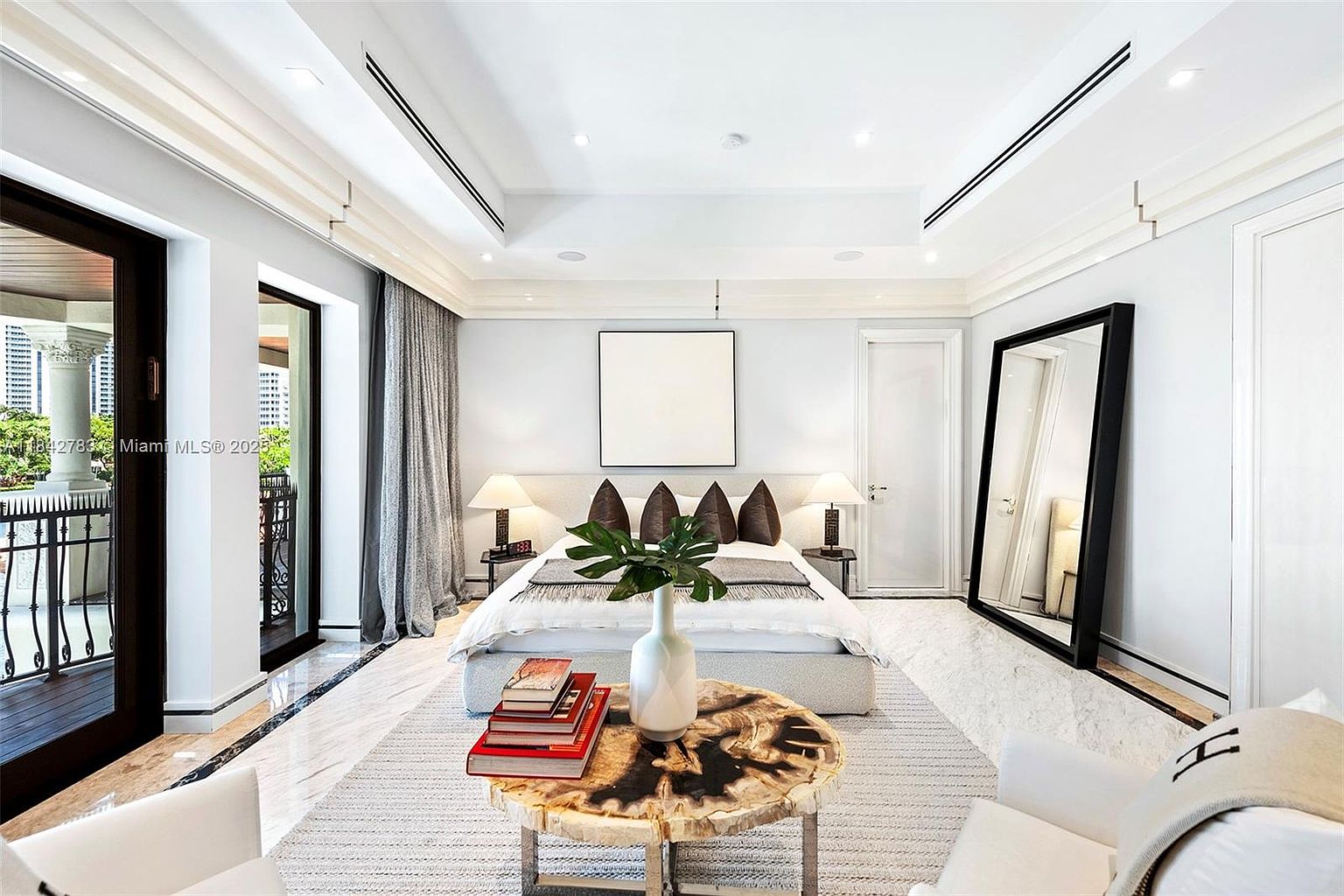 This elegant primary bedroom features a neutral color palette with a central bed flanked by matching lamps and nightstands. A unique, organic-shaped wood coffee table sits in the foreground, while a large, black-framed floor mirror adds a dramatic touch to the right side of the room. The space is brightened by large glass doors that open onto a balcony, creating a seamless transition between the indoor living area and the outdoor space.