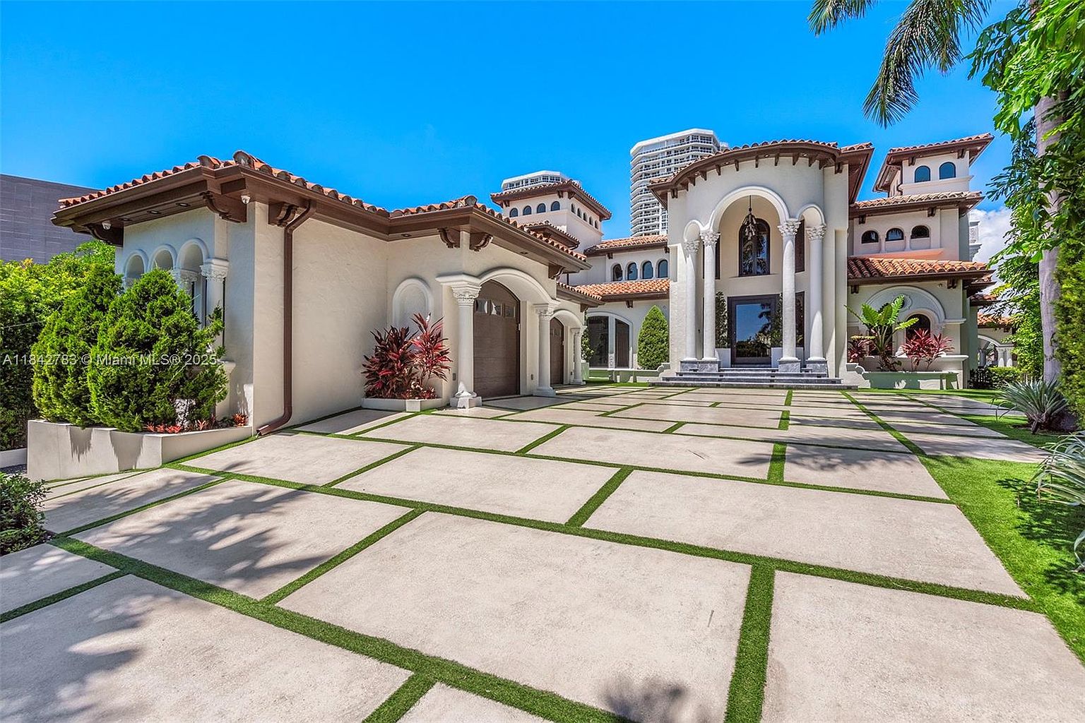 This grand Mediterranean-style estate features a prominent arched entryway supported by classic columns and a multi-level roofline with decorative towers. The expansive driveway is composed of large concrete pavers separated by strips of green turf, leading up to a multi-car garage on the left. Lush landscaping, including manicured shrubs and palm trees, frames the light-colored stucco facade under a clear blue sky.