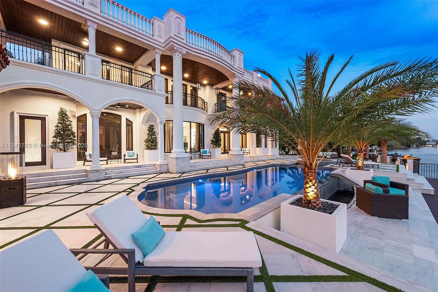 This luxurious waterfront estate features a grand, multi-level exterior with classic architectural columns and a sprawling terrace. A custom-shaped swimming pool serves as the centerpiece of the patio, which is finished with light-toned stone pavers and integrated artificial turf accents. The scene is captured at twilight, highlighting the warm glow of the pool lighting and the elegant, resort-style outdoor living space.