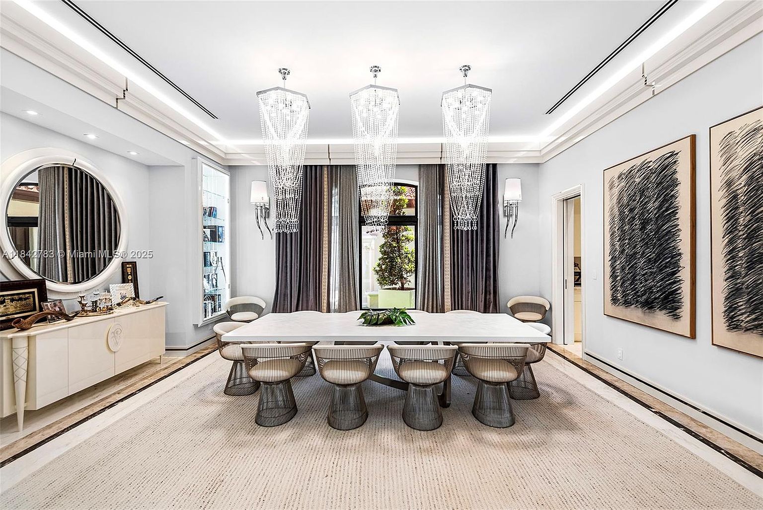 This elegant dining room features a large, rectangular white table surrounded by ten modern, wire-frame chairs with cushioned seats. Three dramatic, tiered crystal chandeliers hang above the table, complemented by wall sconces and a large circular mirror on the left wall. The space is framed by dark, floor-length curtains and two large abstract art pieces, creating a sophisticated and formal atmosphere.