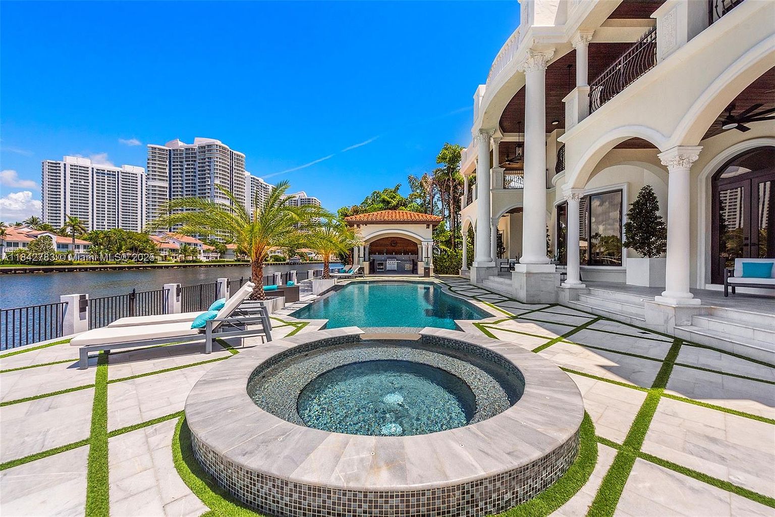 This luxurious waterfront backyard features a circular spa in the foreground that transitions into a rectangular swimming pool. The patio is paved with large stone tiles accented by strips of artificial turf, leading toward a covered outdoor kitchen area and a grand multi-story residence with classical columns. The scene is set against a backdrop of a calm canal and high-rise buildings, creating an upscale, resort-like atmosphere.