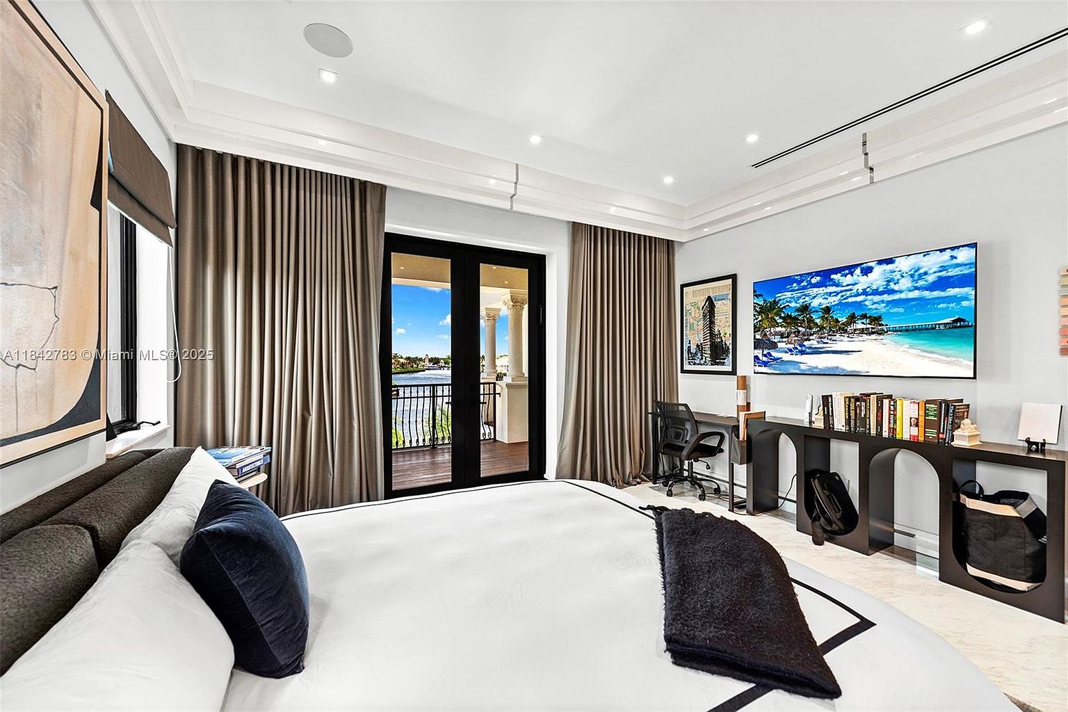 This primary bedroom features a contemporary design with a large bed, a sleek dark console table serving as a desk, and a wall-mounted television displaying a beach scene. Tall glass doors with floor-to-ceiling curtains open onto a private balcony, providing a view of the water and surrounding landscape. The room is finished with light-colored walls and neutral flooring, creating a bright and sophisticated atmosphere.