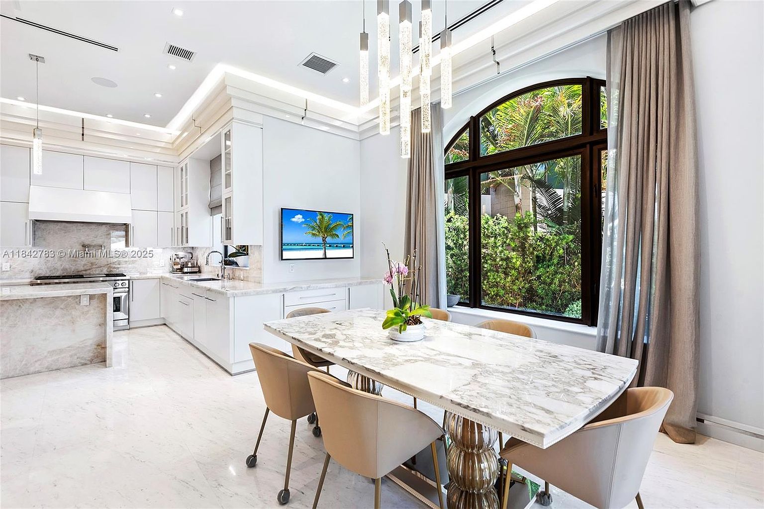 This bright, open-concept kitchen and dining area features sleek white cabinetry and a large stone-topped dining table as the central focal point. A tall, arched window provides a lush view of the outdoors, while modern pendant lighting hangs above the dining space. The room is finished with light-toned stone flooring and a wall-mounted television, creating a sophisticated and airy atmosphere.