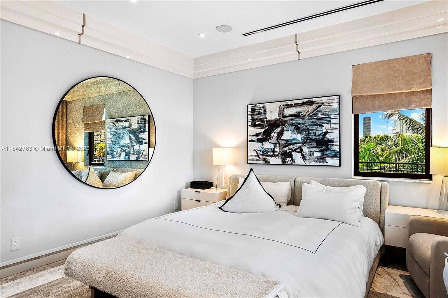 This bright and modern primary bedroom features a plush bed with white linens, accented by a textured bench at the foot. A large, circular mirror hangs on the wall above a nightstand, reflecting the room's clean aesthetic, while a bold abstract painting adds a contemporary focal point above the headboard. A window with a neutral roman shade provides natural light and a view of palm trees and city buildings outside.