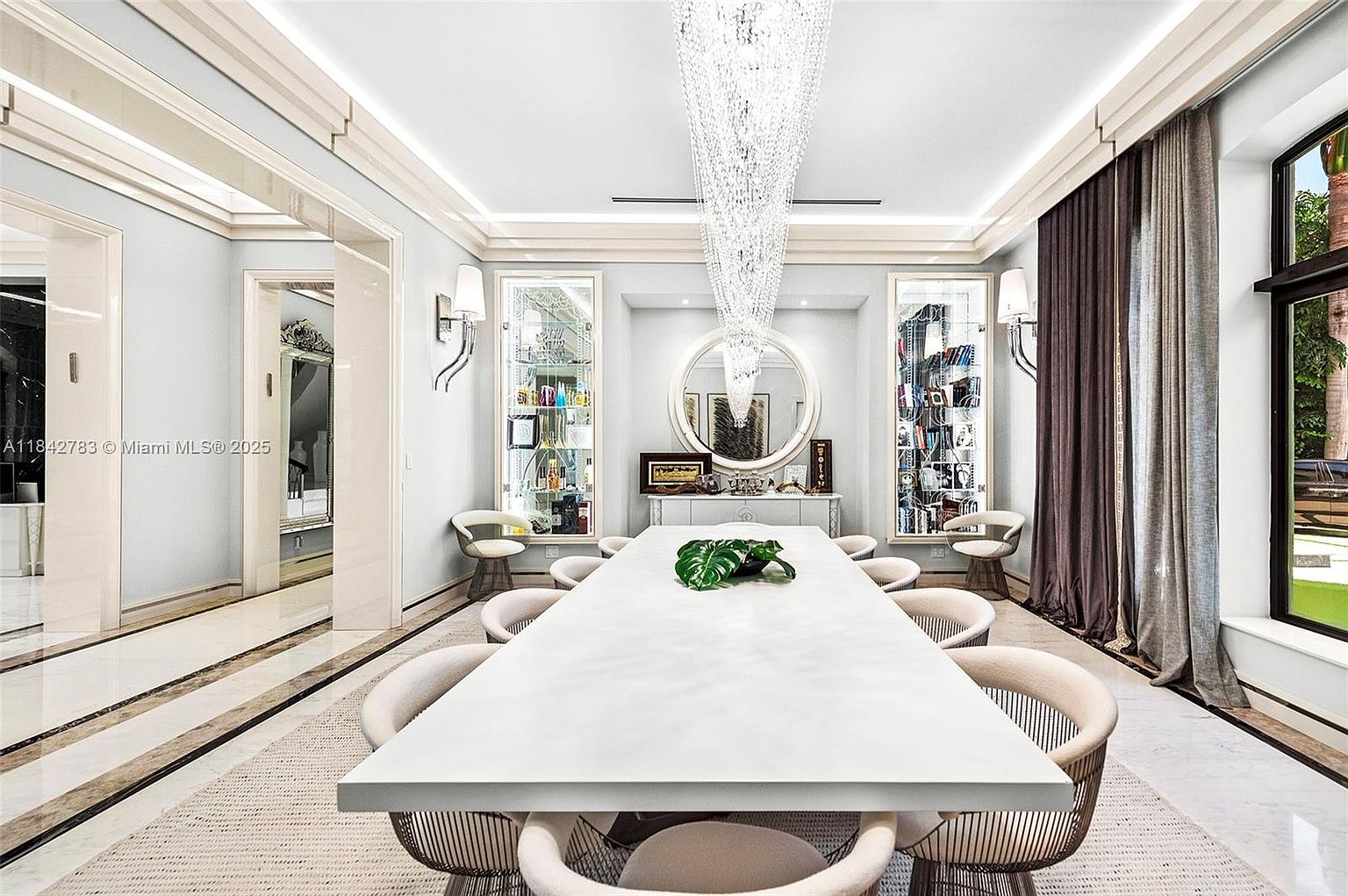 This elegant dining room features a long, minimalist white table surrounded by modern, curved-back chairs set atop a textured area rug. A dramatic, multi-tiered crystal chandelier serves as the focal point above the table, while symmetrical built-in display cabinets flank a large circular mirror on the back wall. The space is framed by tall windows with floor-length curtains and polished stone flooring, creating a sophisticated and airy atmosphere.
