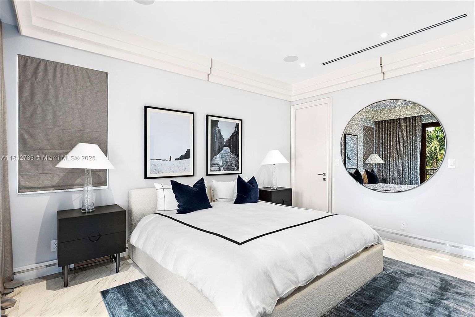 This elegant primary bedroom features a neutral color palette with a plush, light-toned bed as the central focal point, accented by dark navy pillows and a crisp white duvet. The room is symmetrically arranged with matching black nightstands, table lamps, and framed black-and-white wall art, while a large circular mirror adds a modern decorative touch. The space is finished with light-colored flooring and a textured area rug, creating a sophisticated and serene atmosphere.