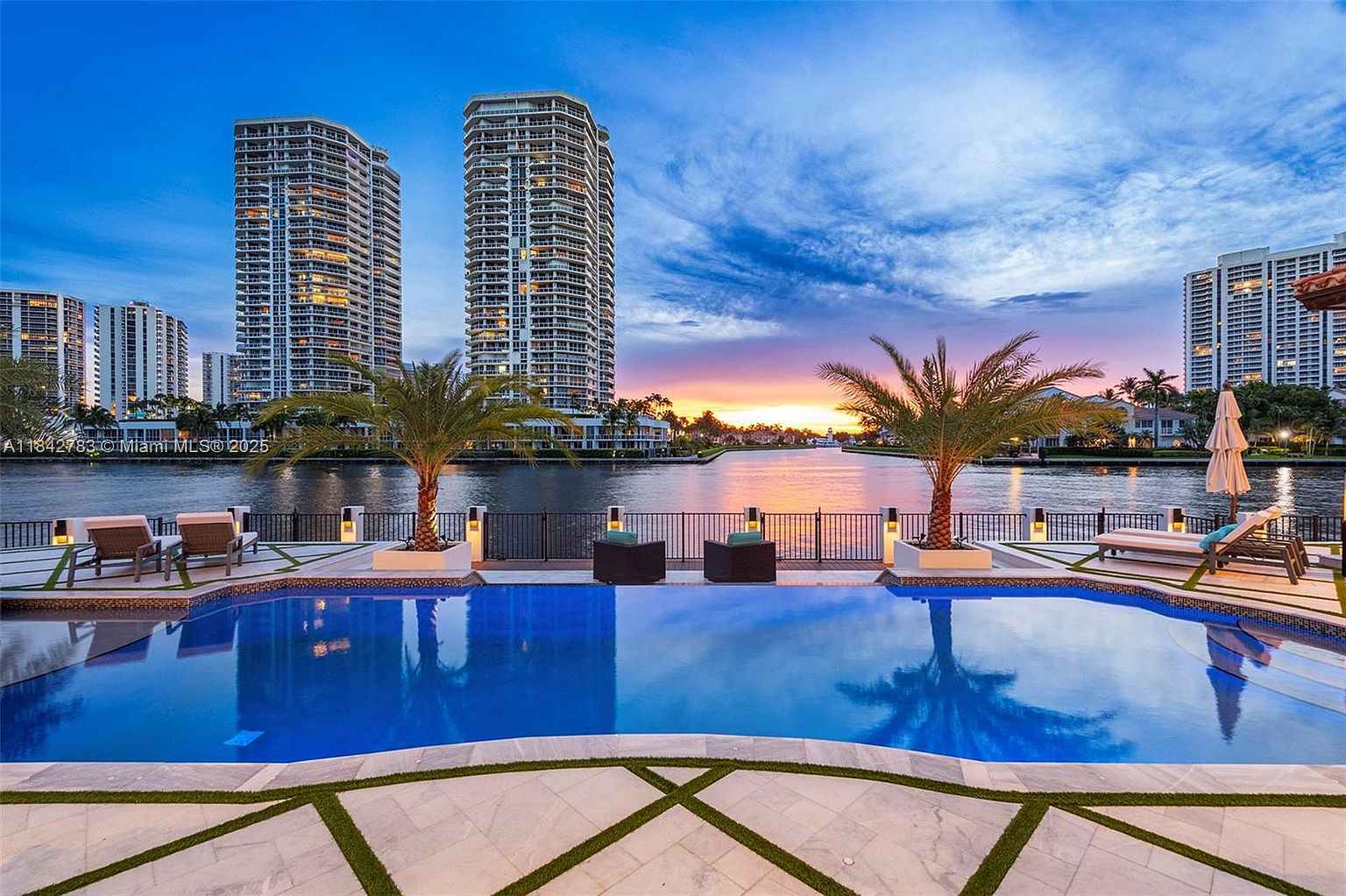 This stunning waterfront pool area offers a panoramic view of a tranquil canal and high-rise city skyline at sunset. The scene features a pristine blue swimming pool framed by elegant stone decking, manicured palm trees, and comfortable outdoor lounge furniture. The composition captures a serene, luxurious atmosphere, perfectly highlighting the seamless transition between the private residence and the scenic waterway.