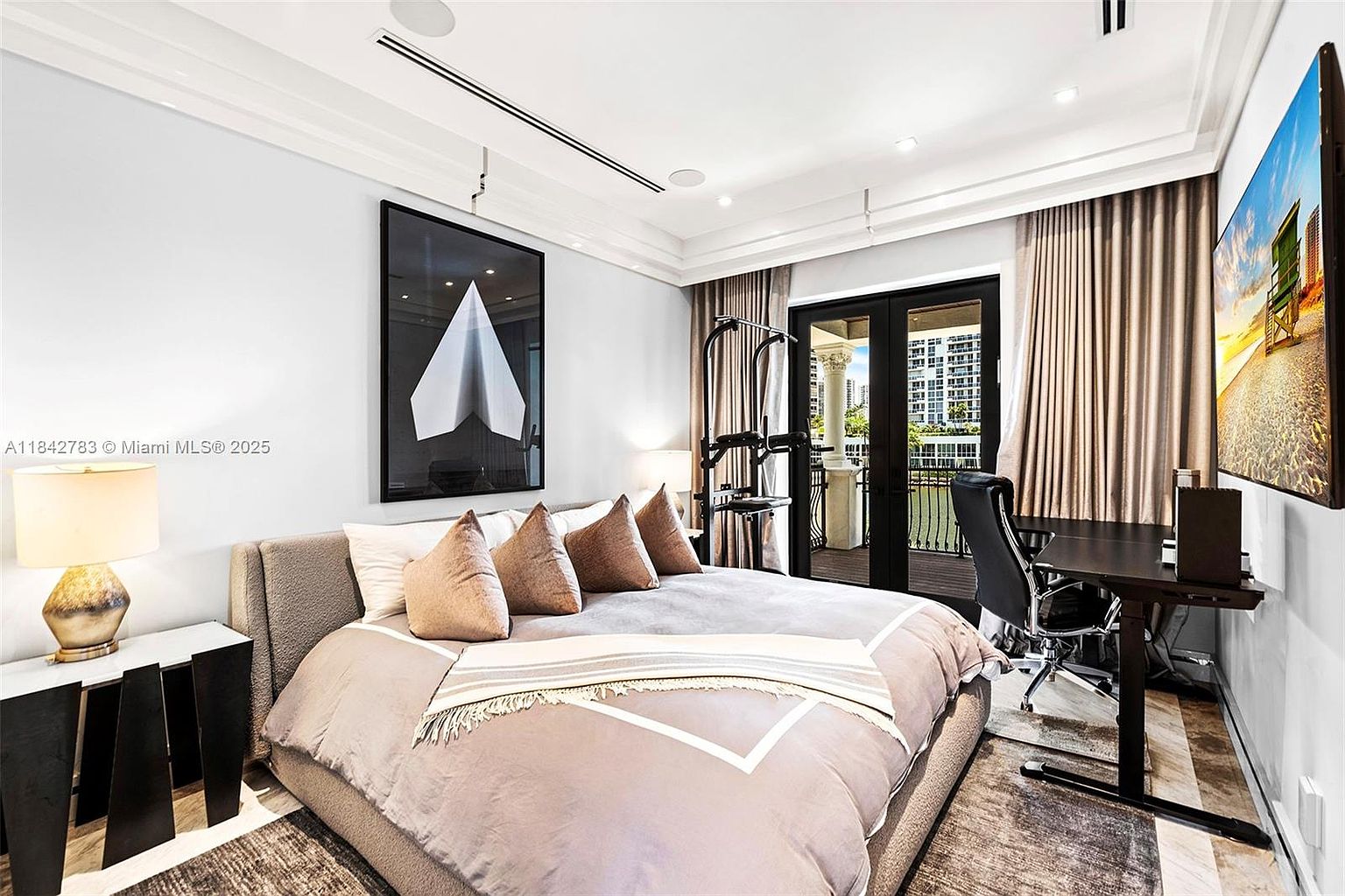 This modern bedroom features a neutral color palette with a plush upholstered bed adorned with several decorative pillows. To the right, a sleek desk setup with an ergonomic chair is positioned near large glass doors that open to a balcony, while a home gym station is tucked into the corner. The room is accented by a large framed piece of abstract art on the wall and a bedside lamp, creating a sophisticated and multi-functional living space.