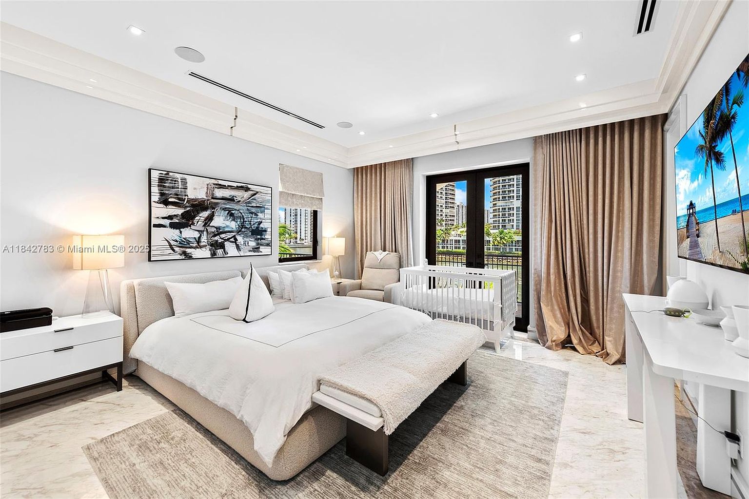 This spacious primary bedroom features a neutral color palette with a large bed centered against a light-toned wall adorned with abstract art. The room includes a crib, a comfortable armchair, and a sleek white desk, all set upon a textured area rug over light stone flooring. Large glass doors with floor-to-ceiling curtains provide access to an outdoor view, creating a bright and serene atmosphere.