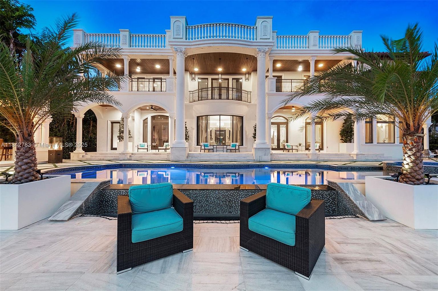This grand, multi-story estate features a symmetrical neoclassical design with white columns, expansive balconies, and a large swimming pool in the foreground. Two modern, dark-wicker armchairs with vibrant turquoise cushions sit on the stone patio, creating a luxurious lounge area overlooking the water. The scene is captured at dusk, with warm lighting illuminating the home's architectural details and reflecting off the pool's surface.