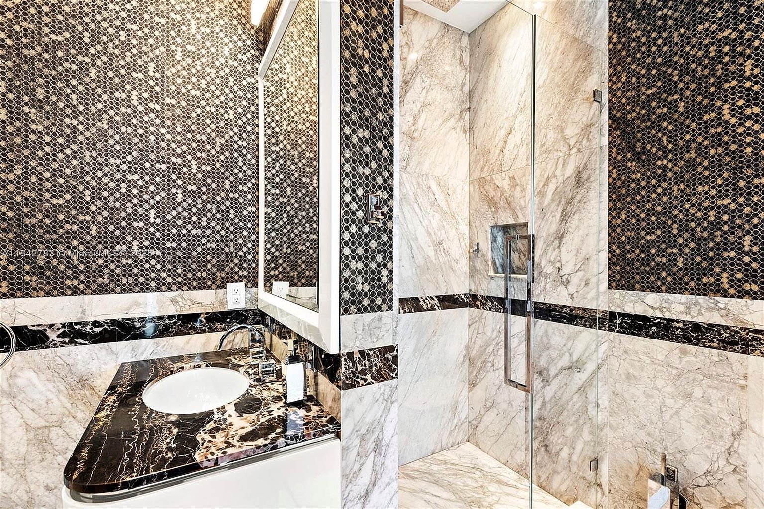 This luxurious bathroom features a striking contrast between dark, patterned stone surfaces and light, veined stone wall tiles. The vanity is topped with a bold, dark stone counter and includes a sleek, modern sink, while the adjacent walk-in shower is enclosed by a glass panel and features a recessed niche. The space is accented with intricate, dark mosaic wall tiles that add a sophisticated, high-end texture to the room.