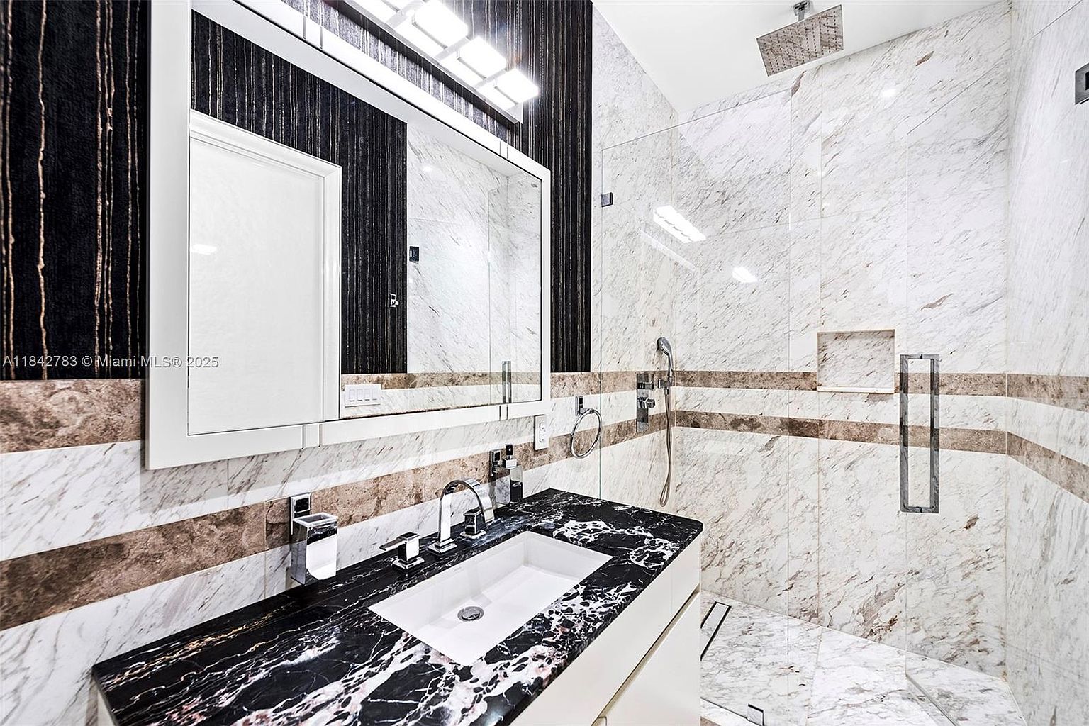 This modern bathroom features a striking vanity with a dark, heavily veined stone countertop and a white undermount sink. A large, rectangular mirror is mounted against a dark, textured accent wall, while the adjacent shower area is enclosed by glass and finished with light-toned, veined stone tiles. The space is illuminated by a sleek, multi-bulb light fixture above the mirror and a rainfall showerhead, creating a sophisticated and high-contrast aesthetic.