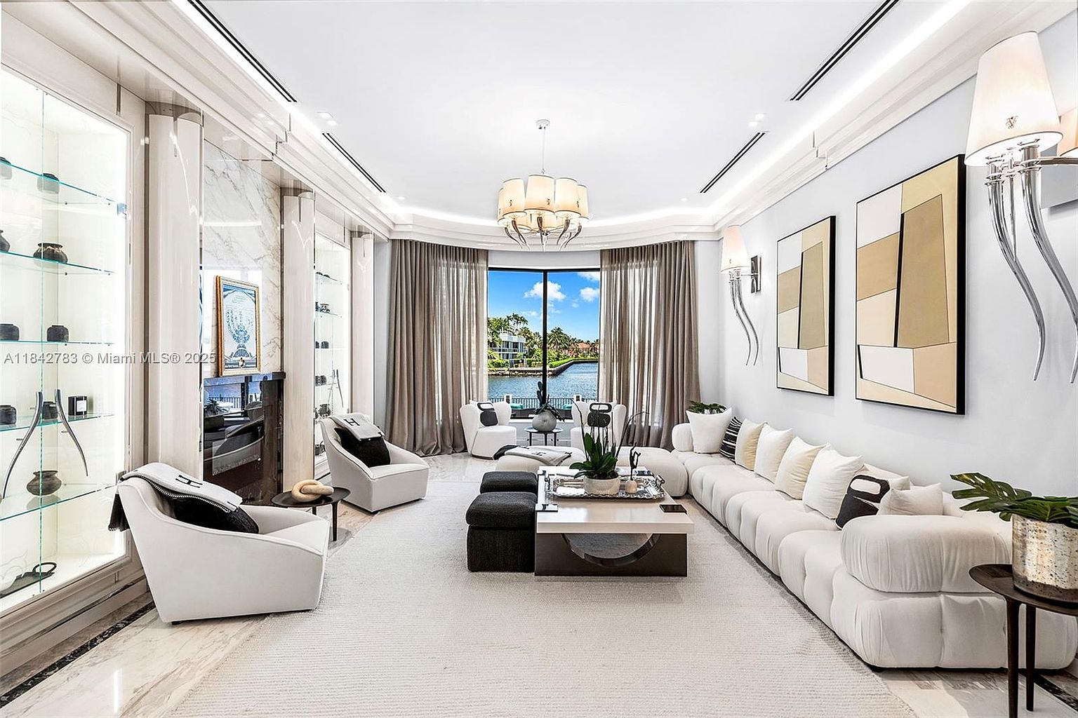 This sophisticated living room features a bright, open-concept design with a large white sectional sofa and matching armchairs arranged around a central coffee table. The space is anchored by a tall stone-clad fireplace surround and illuminated by a decorative chandelier, while large windows provide a clear view of the water outside. The room is accented with modern artwork and glass display shelving, creating a luxurious and airy atmosphere.