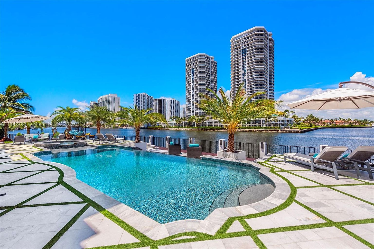 This luxurious waterfront pool area features a custom-shaped swimming pool with a built-in spa, surrounded by light-toned stone pavers with integrated grass accents. The deck is furnished with modern lounge chairs and a large umbrella, offering a clear view of the calm canal and the towering high-rise buildings in the background. The scene is captured from a low-angle perspective, emphasizing the expansive outdoor living space and the serene, sunny atmosphere.