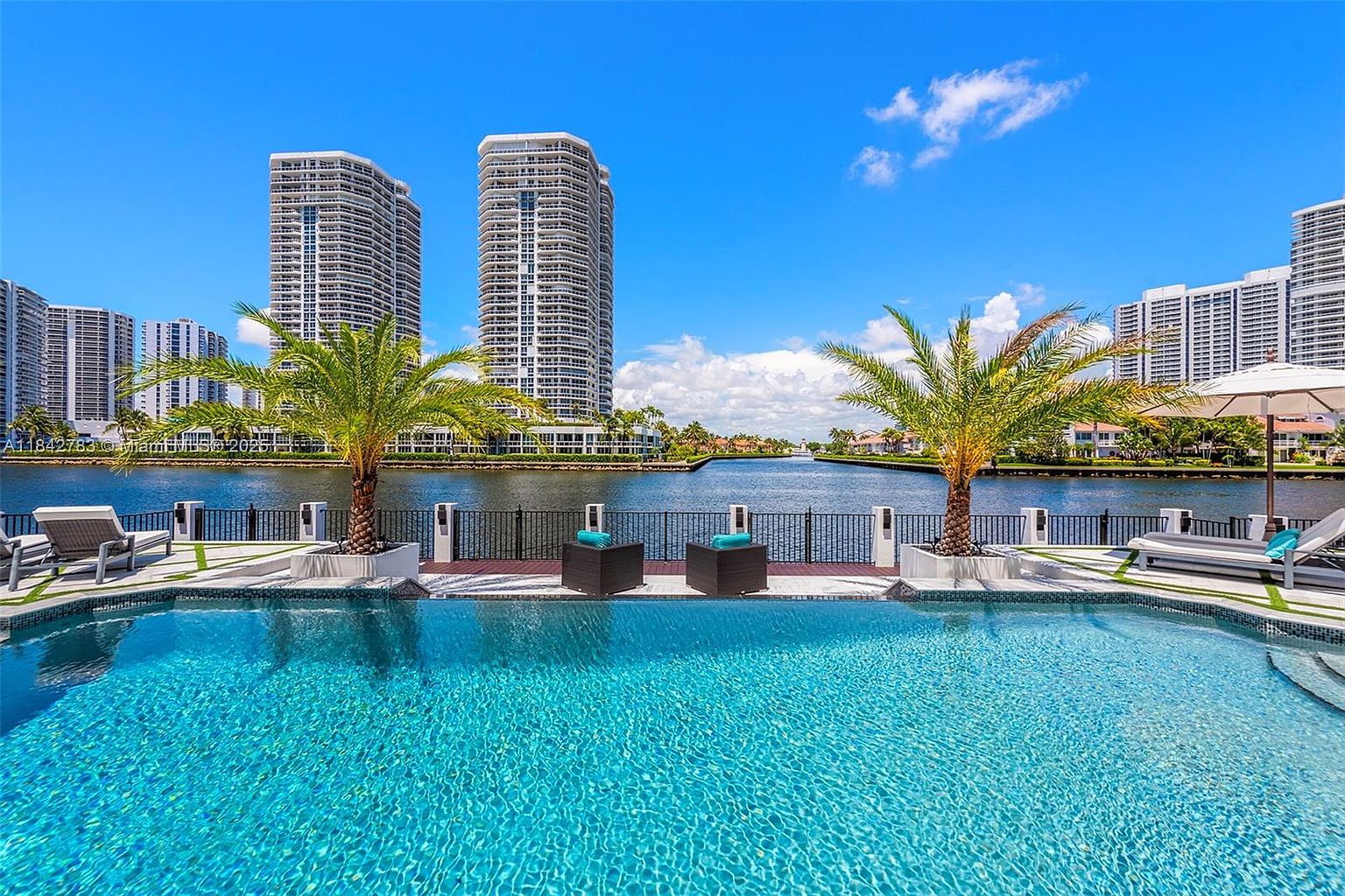 This stunning waterfront pool area offers a luxurious outdoor living experience with a clear view of high-rise buildings across the canal. The scene features a sparkling blue swimming pool in the foreground, flanked by two palm trees and modern lounge chairs on a paved deck. A black metal fence separates the pool area from the water, creating a serene and sophisticated atmosphere perfect for relaxation.