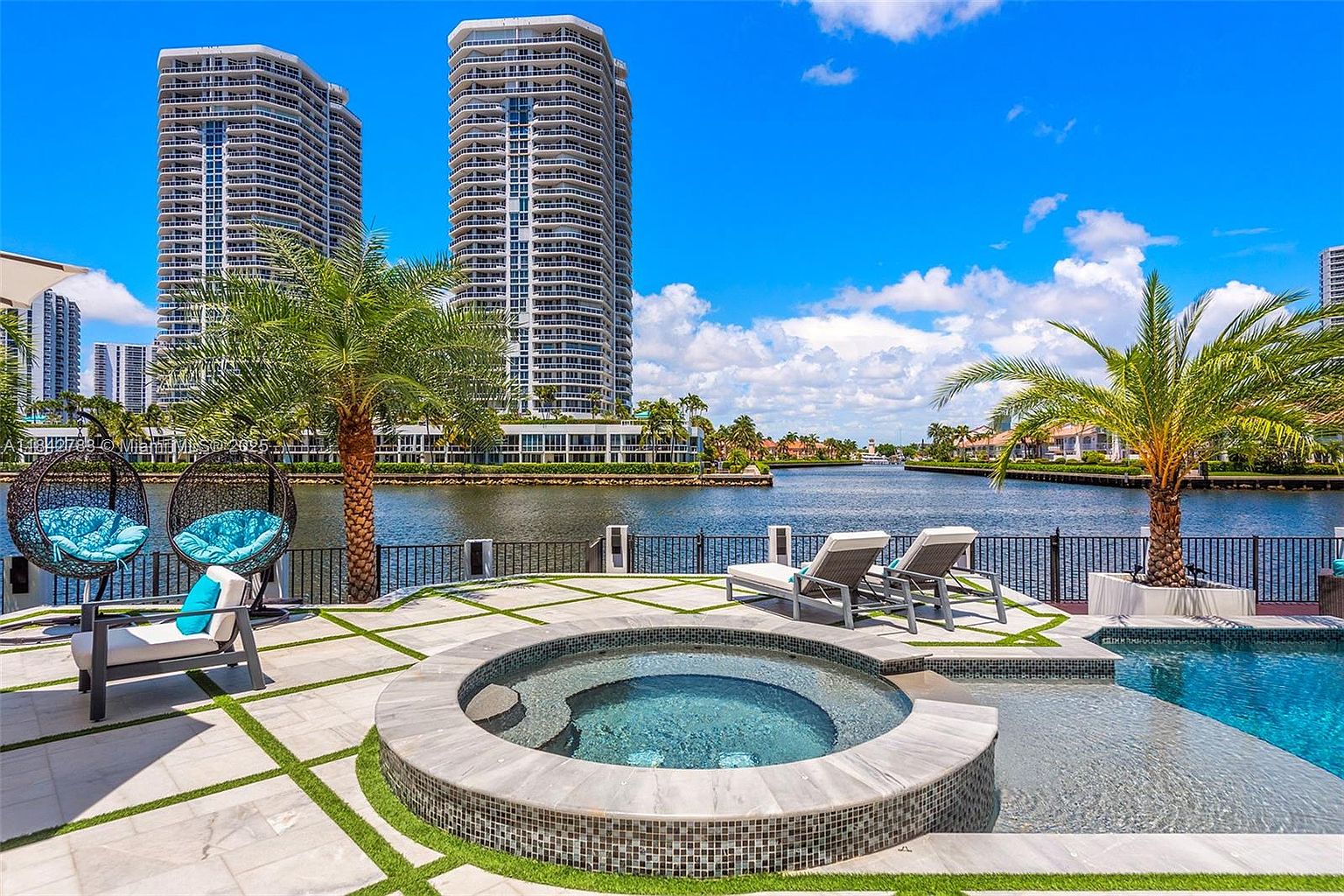 This luxurious waterfront patio features a circular tiled spa and a portion of a swimming pool, set against a backdrop of a calm canal and high-rise residential towers. The outdoor living space is furnished with two modern lounge chairs, a comfortable armchair with a bright blue cushion, and two hanging egg chairs, all arranged on a paved deck with integrated grass accents. The scene captures a serene, upscale tropical lifestyle with clear blue skies and lush palm trees framing the view.