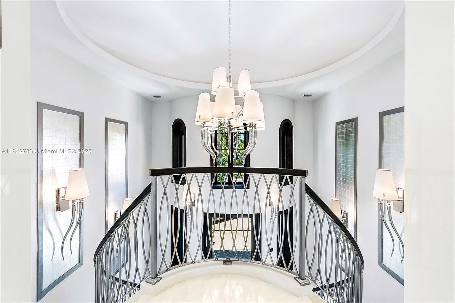 This elegant upper-level landing features a curved balcony with a decorative metal railing that overlooks the space below. A grand, multi-tiered chandelier serves as the focal point, suspended from a recessed circular ceiling detail. The area is framed by symmetrical wall sconces and tall, narrow windows, creating a bright and sophisticated atmosphere.
