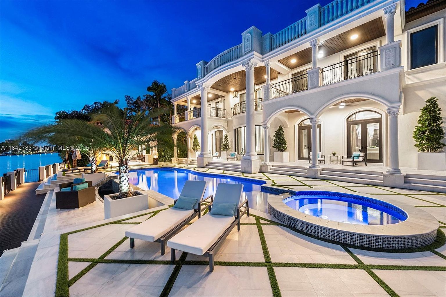 This luxurious waterfront estate features a grand two-story rear facade with classic architectural columns and multiple balconies overlooking a resort-style pool and spa area. The expansive stone patio is accented with geometric grass inlays, creating a sophisticated outdoor living space perfect for entertaining. The scene is captured at twilight, highlighting the warm glow of the home's lighting against the serene backdrop of the water.