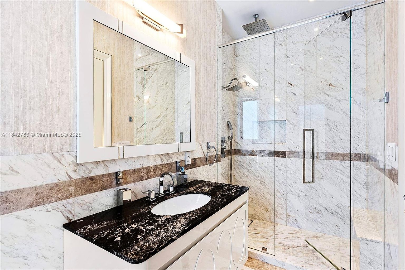 This modern bathroom features a striking contrast between a dark, veined stone vanity top and light-toned, patterned wall tiles. A glass-enclosed shower stall with a rainfall showerhead and a handheld sprayer occupies the right side of the space, while a rectangular mirror hangs above the vanity. The room is illuminated by a sleek, linear light fixture, creating a clean and sophisticated aesthetic.