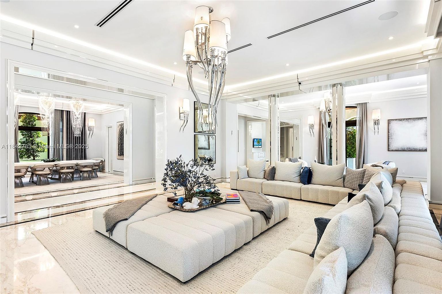 This expansive, light-filled living room features a neutral color palette with a large, modular sectional sofa and a central oversized ottoman. A striking, multi-tiered crystal chandelier serves as the focal point, while mirrored wall panels enhance the sense of space and reflect the adjacent dining area. The room is characterized by its high ceilings, recessed lighting, and a sophisticated, contemporary aesthetic.