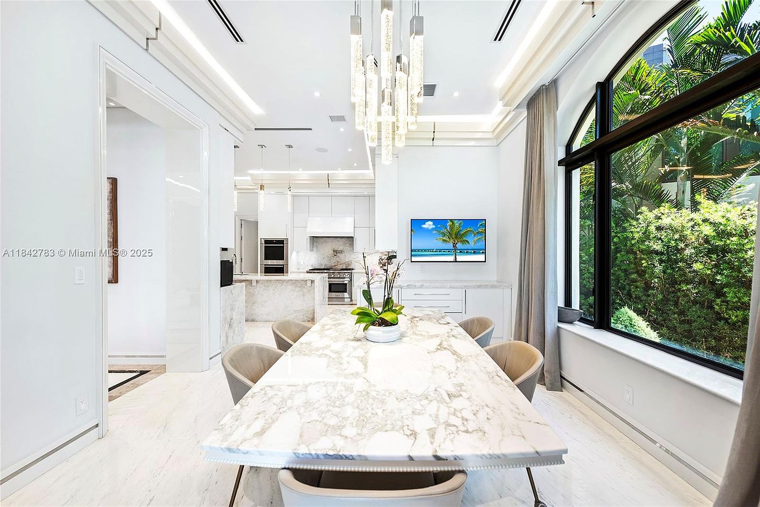 This elegant dining area features a large stone-topped table surrounded by upholstered chairs, positioned beneath a striking multi-pendant light fixture. Large windows provide a lush view of the outdoor greenery, while the open-concept layout flows seamlessly into a bright, modern kitchen in the background. The space is defined by its clean white aesthetic, polished light-toned flooring, and a wall-mounted television, creating a sophisticated and airy atmosphere.