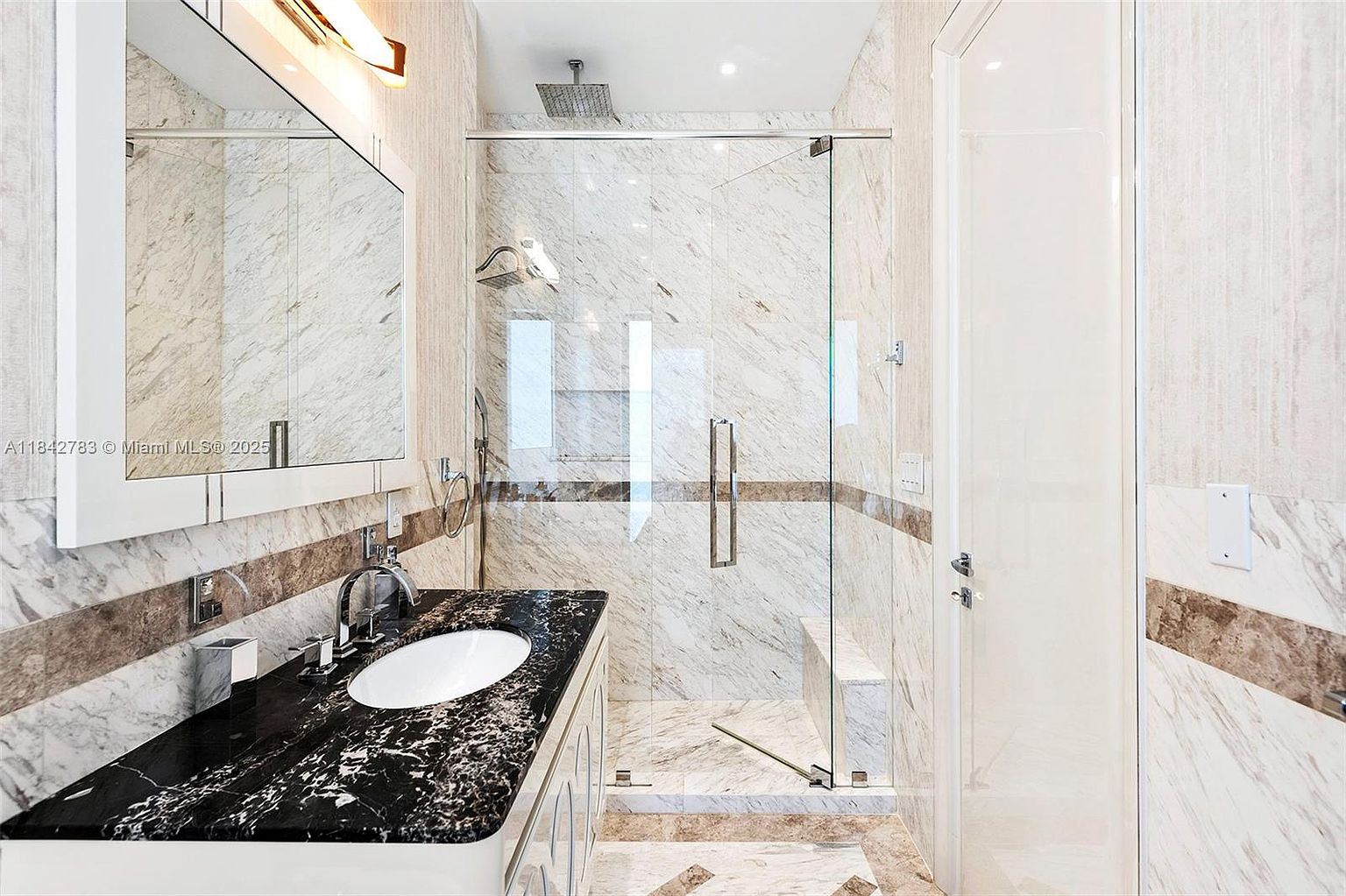 This elegant bathroom features a vanity with a dark, heavily veined stone countertop and a white undermount sink, complemented by a large wall-mounted mirror. To the right, a glass-enclosed shower stall is finished with light-toned stone wall tiles and includes a built-in bench. The space is illuminated by a warm light fixture above the mirror, creating a sophisticated and polished atmosphere.