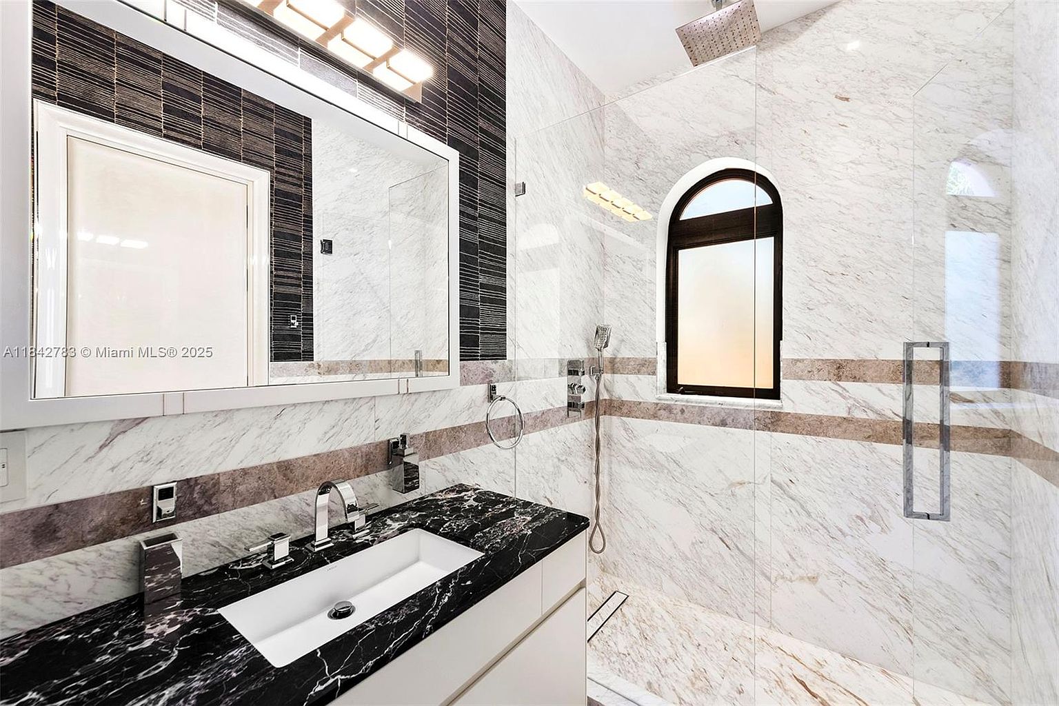 This modern bathroom features a striking contrast between a black stone vanity countertop and light-toned, veined stone wall tiles. A large rectangular mirror sits above the vanity, complemented by a sleek faucet and a walk-in shower area enclosed by a glass partition. The space is illuminated by a contemporary light fixture above the mirror and a window with an arched top, creating a bright and sophisticated atmosphere.