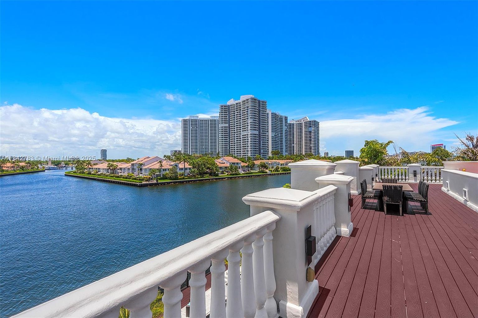 This elevated balcony features a dark-toned deck surface and a classic white balustrade railing, providing a clear vantage point over a calm waterway. A dining set with woven chairs sits on the deck, while the background showcases a scenic view of residential homes and high-rise apartment buildings under a bright blue sky. The perspective is wide and cinematic, emphasizing the expansive outdoor living space and the waterfront location.
