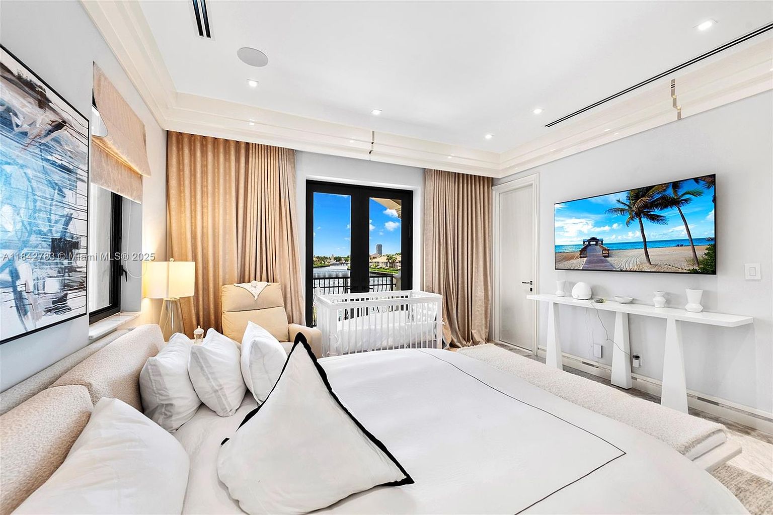 This primary bedroom features a neutral color palette with a large, plush bed as the focal point, complemented by a white crib and a comfortable armchair near the window. A wall-mounted television sits above a sleek, minimalist console table, while floor-to-ceiling curtains frame the glass doors that lead to a balcony with water views. The room is illuminated by recessed lighting and a bedside lamp, creating a serene and sophisticated atmosphere.