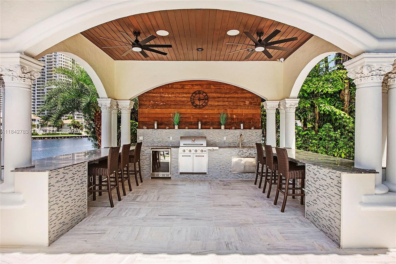 This luxurious outdoor kitchen and bar area is sheltered under a vaulted, wood-paneled ceiling with dual ceiling fans. The space features a built-in grill, a stainless steel beverage refrigerator, and a sink, all set against a rustic wood-paneled wall with a decorative clock. The bar counter is lined with wicker-style stools, and the entire structure is supported by classic columns, offering a scenic view of the waterfront.