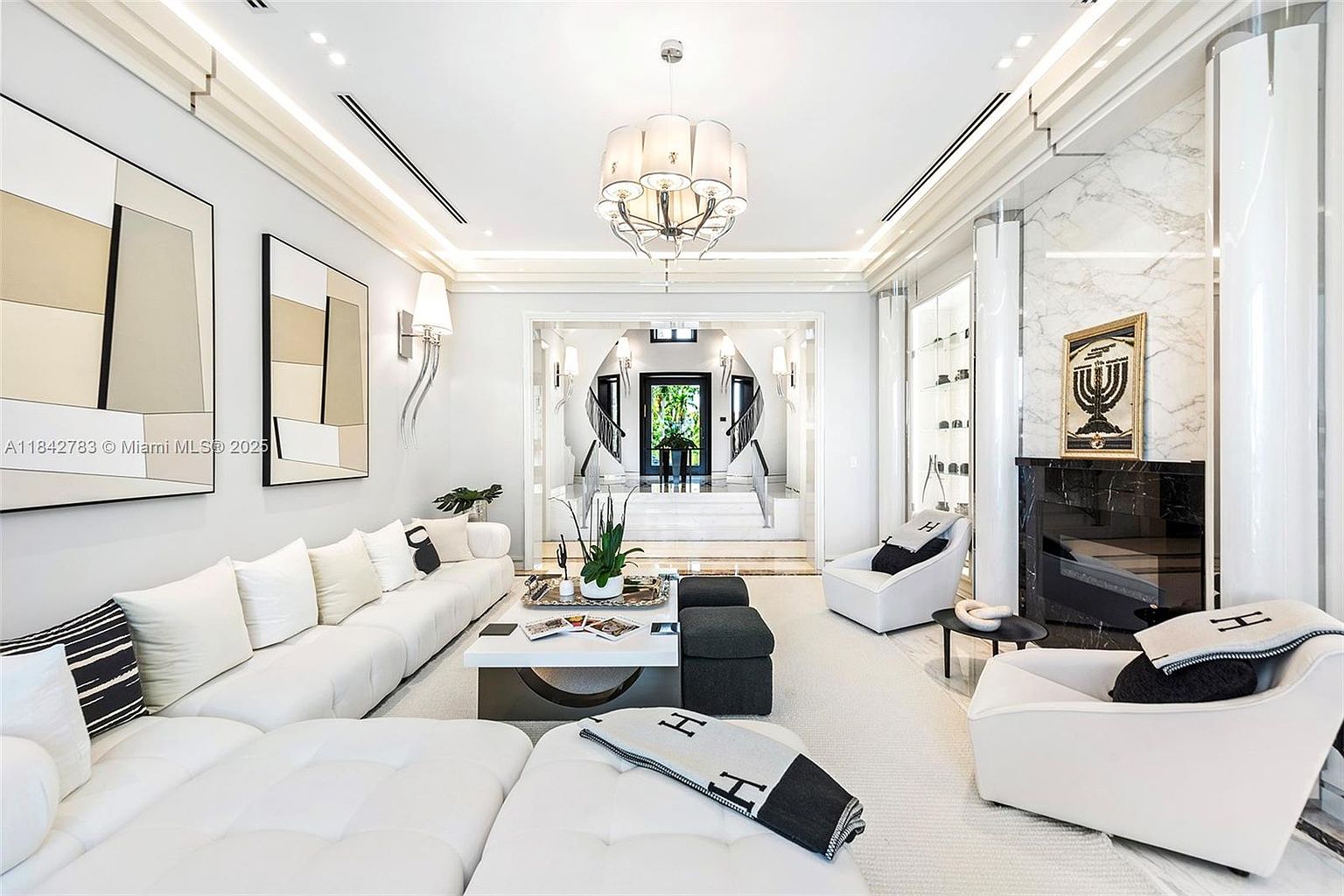 This elegant, monochromatic living room features a large, plush white sectional sofa and matching armchairs arranged around a central coffee table. A striking black stone fireplace surround serves as a focal point, complemented by a tall, white stone-clad wall and a decorative framed piece of art. The space is illuminated by a sophisticated multi-tiered chandelier and recessed lighting, creating a bright, luxurious atmosphere that flows toward a grand entryway with a double staircase.