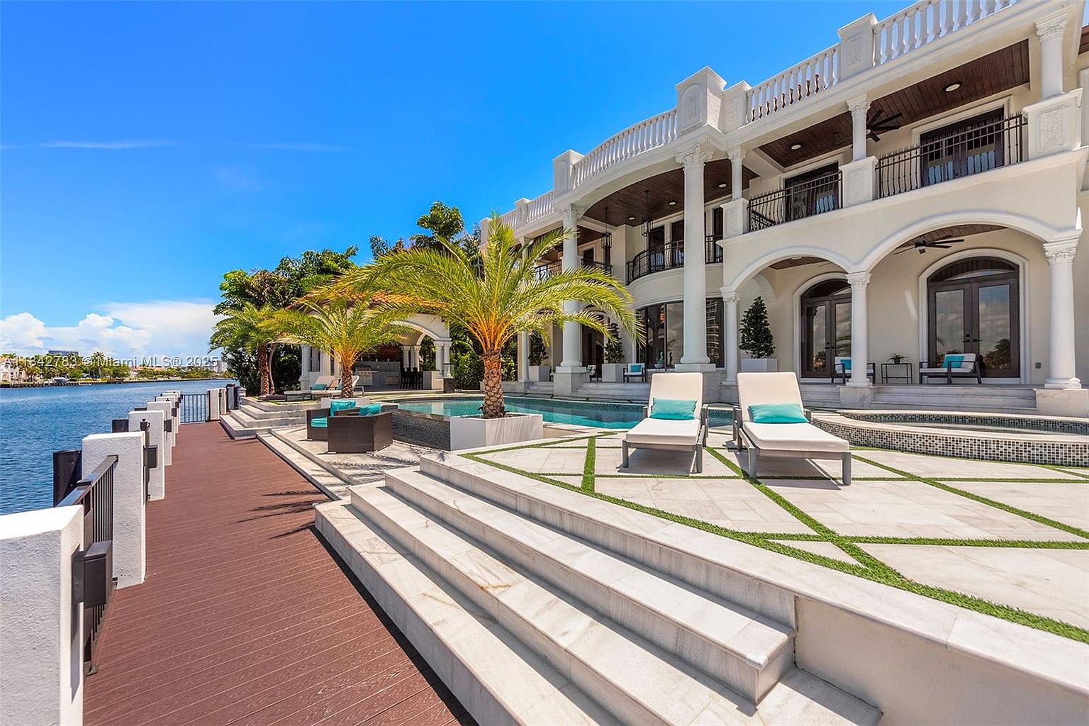 This luxurious waterfront estate features a sprawling stone patio with a custom pool and spa, set against a backdrop of a grand, multi-story white residence. The scene includes a private wooden dock extending over the water, manicured palm trees, and elegant lounge chairs arranged for relaxation. The architecture showcases classic columns and expansive covered balconies, creating a sophisticated and serene outdoor living space.