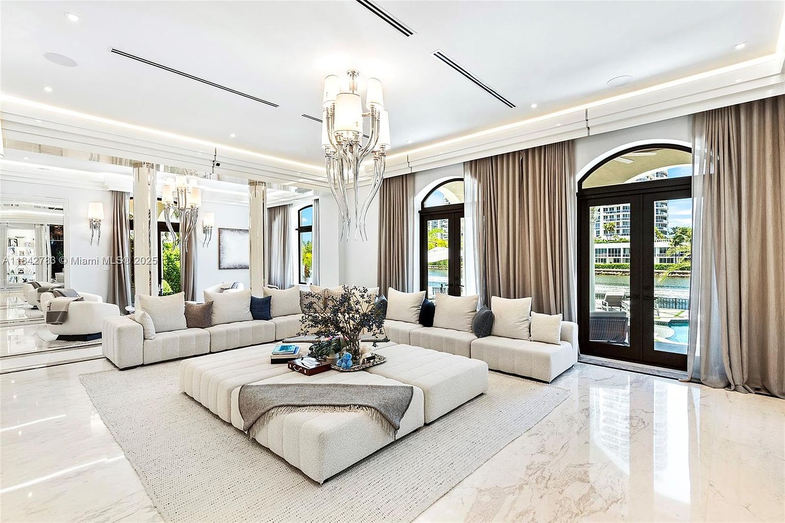 This expansive, light-filled living room features a large, modular sectional sofa arranged around a massive, square upholstered ottoman. The space is defined by polished stone flooring, a dramatic tiered crystal chandelier, and tall glass doors that provide a clear view of the waterfront exterior. The neutral color palette and mirrored wall accents create an atmosphere of sophisticated, modern luxury.