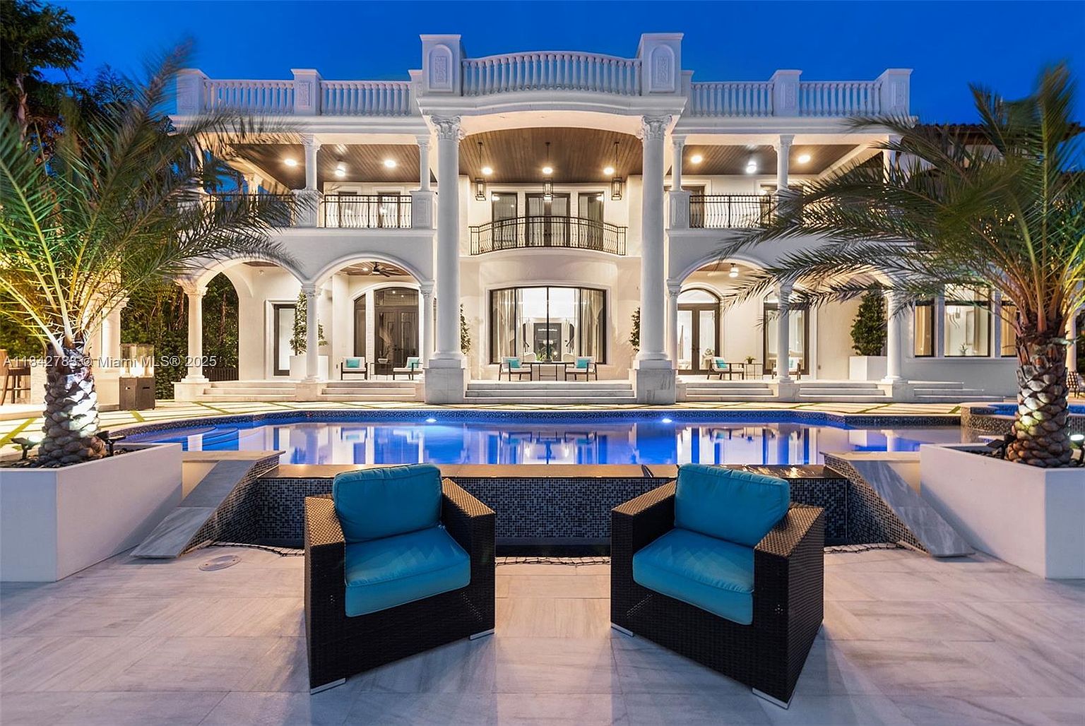This grand, multi-story estate features a symmetrical neoclassical design with prominent white columns and a sprawling balcony overlooking a custom swimming pool. The rear facade is illuminated at dusk, highlighting the architectural details and the inviting patio area furnished with two modern lounge chairs. The composition captures a luxurious outdoor living space, emphasizing the seamless transition between the elegant residence and the tranquil pool deck.