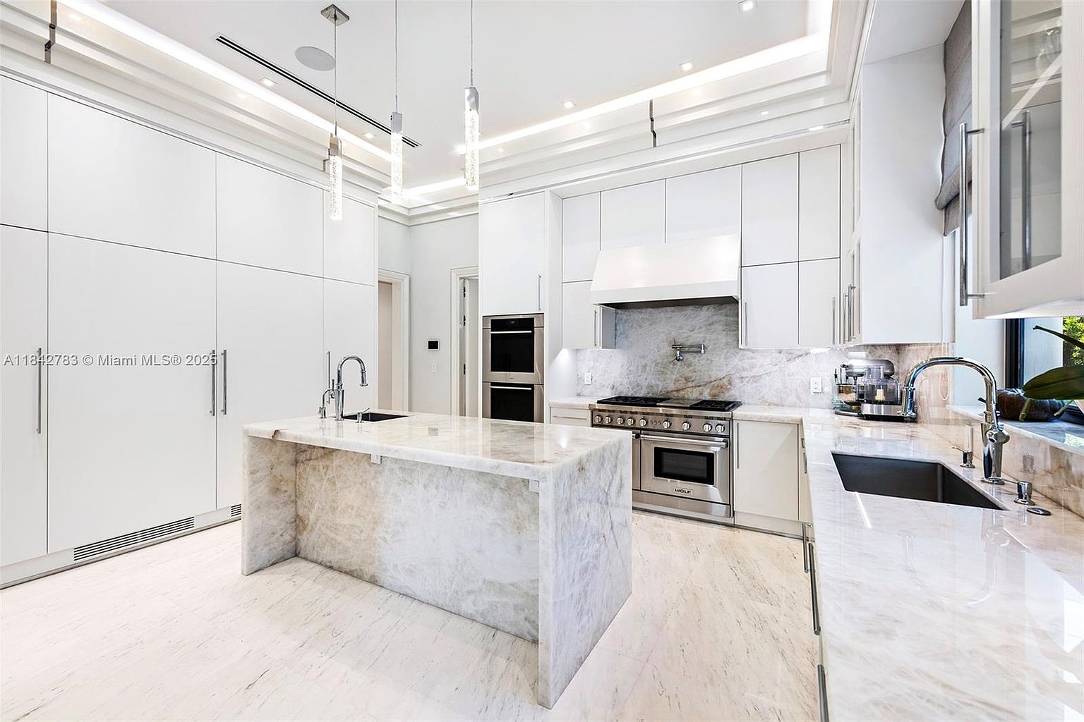 This bright, modern kitchen features sleek white cabinetry and a large central island topped with light-toned stone. The space is equipped with high-end stainless steel appliances, including a professional-grade range and built-in wall ovens. Pendant lights hang above the island, while recessed lighting and a tray ceiling design add depth to the clean, minimalist aesthetic.