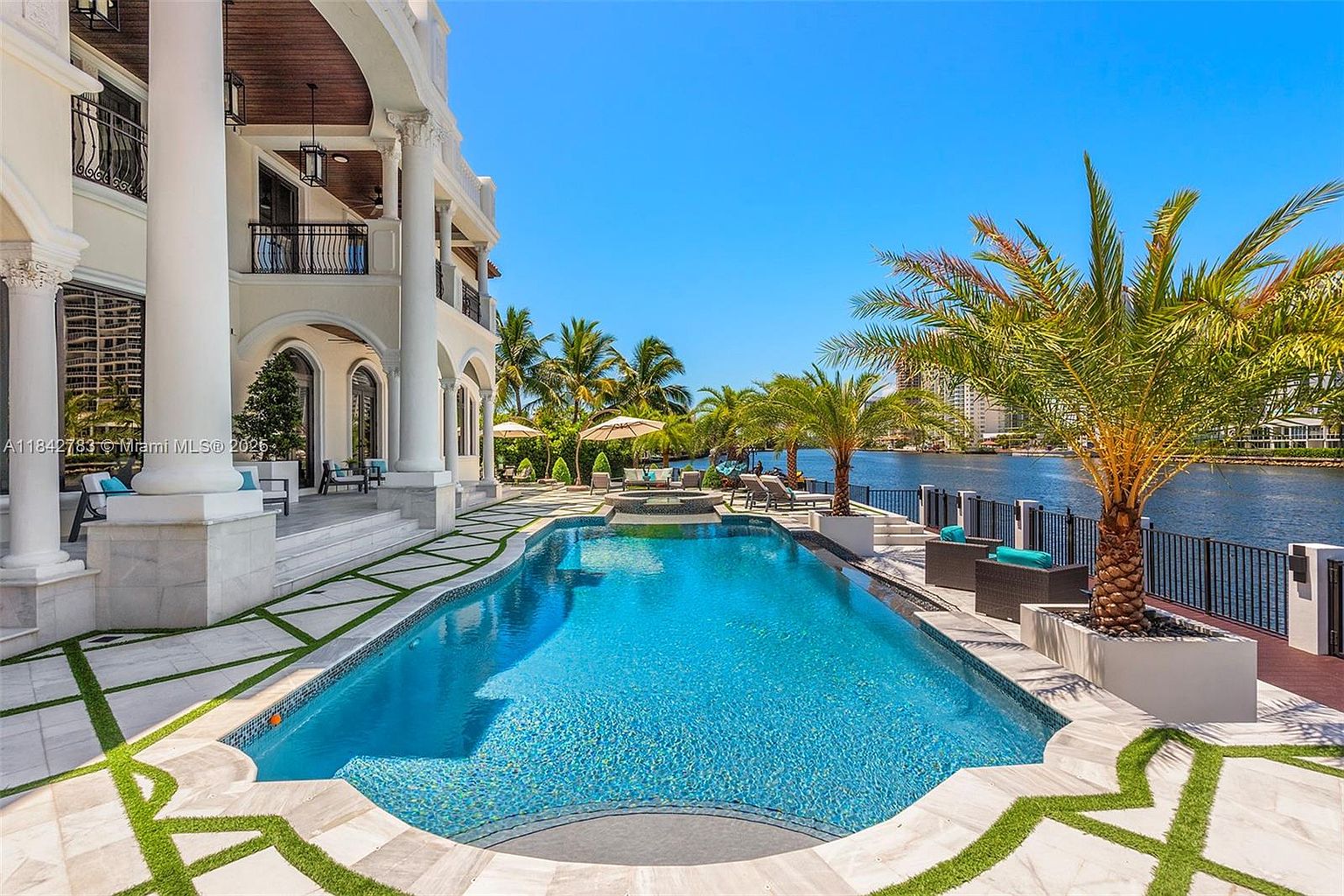 This luxurious waterfront estate features a sprawling pool and spa area overlooking a calm canal. The architecture showcases grand white columns and a covered patio, while the pool deck is finished with light-toned stone pavers accented by geometric grass inlays. A mature palm tree stands prominently near the water's edge, creating a serene and upscale tropical atmosphere.